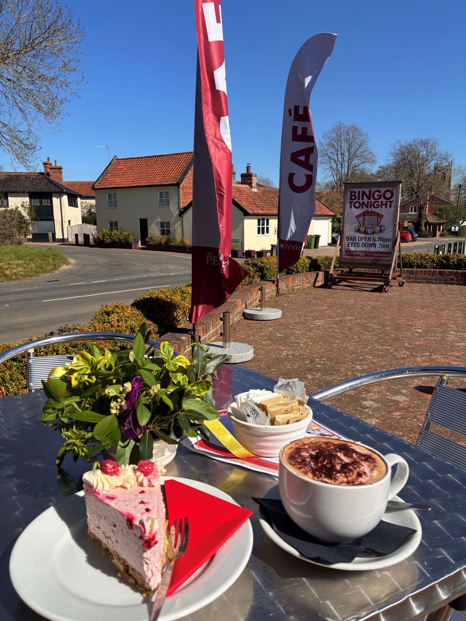 Outdoor seating area at The Pennoyer Centre in Pulham St Mary with café tables, flowers, cakes and promotional flags outside the historic building.