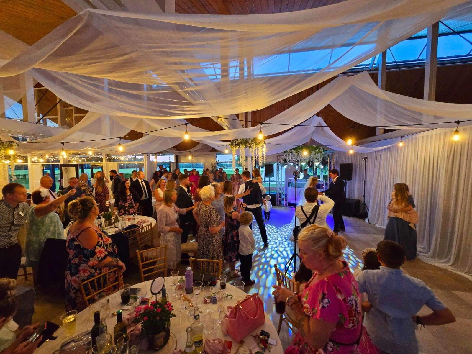 Wedding reception in a decorated pavilion with draped fabric, festoon lighting, dining tables and guests gathered around the dance floor.