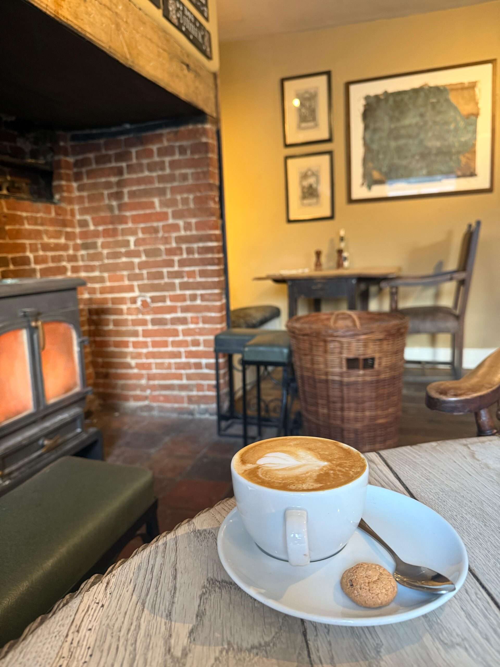 A cosy seating area inside The White Hart with a wood-burning stove, exposed brickwork, rustic furniture and a freshly made coffee served on a wooden table.