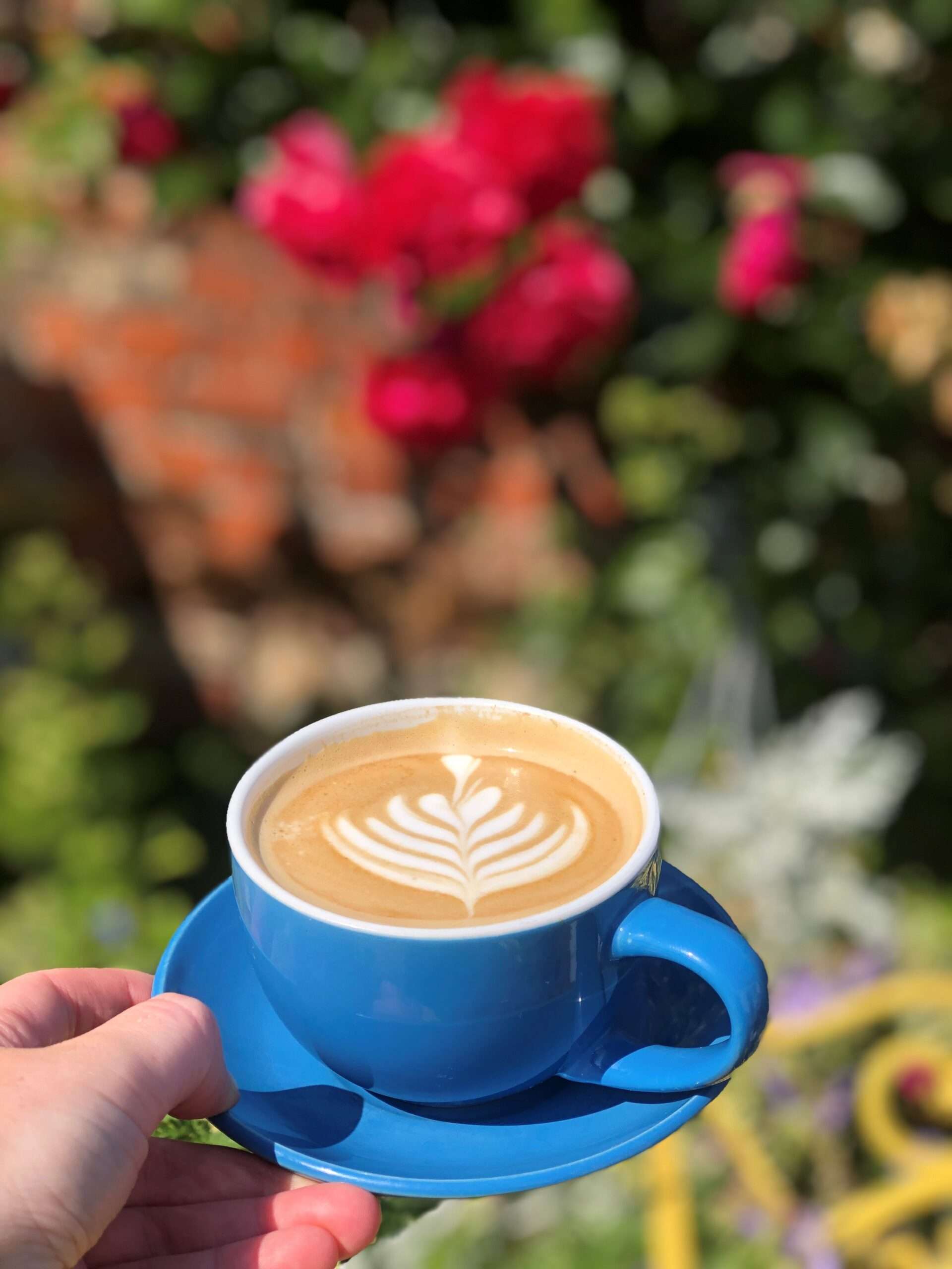 Close-up of a latte with leaf-shaped latte art on a wooden table, with colourful flowers softly blurred in the background.
