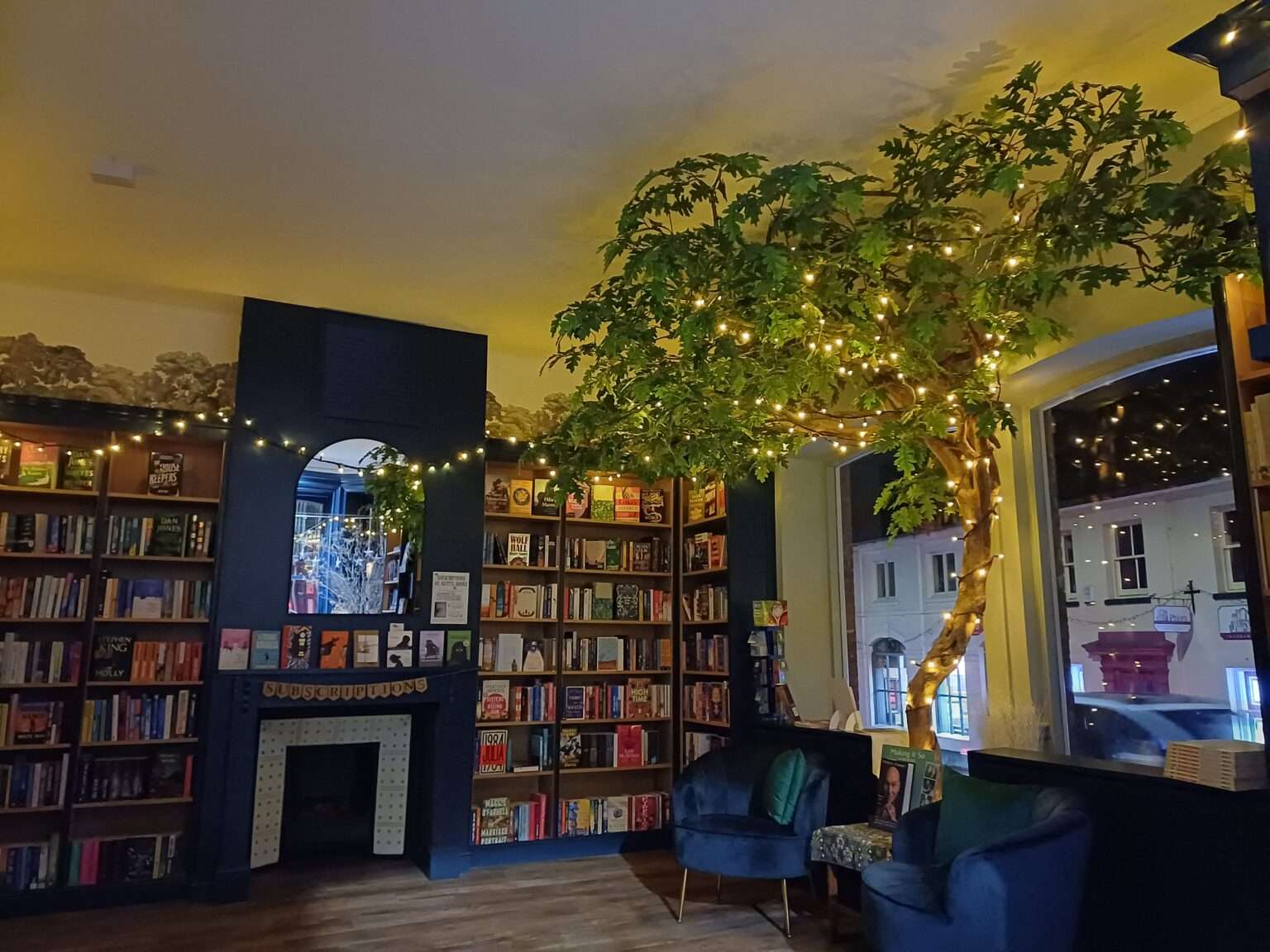 Cosy evening view inside Kett’s Books, with illuminated indoor tree, armchairs, fireplace and shelves of books.
