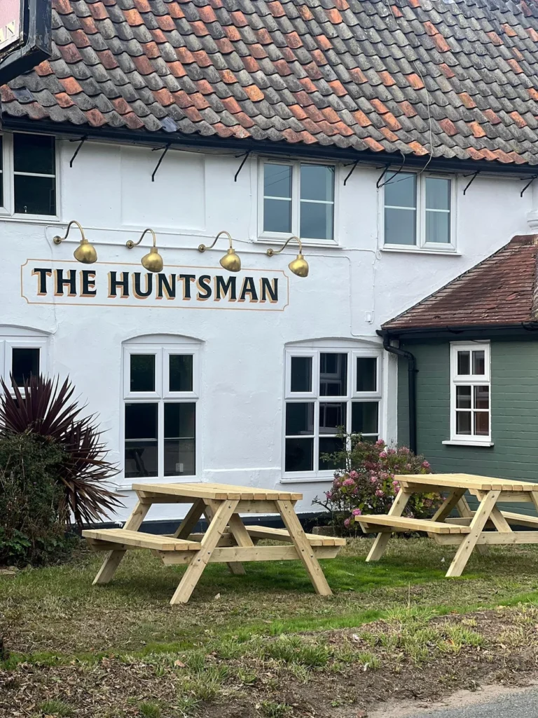 Exterior view of The Huntsman in Strumpshaw, showing the traditional pub building, outdoor seating area, and village setting.